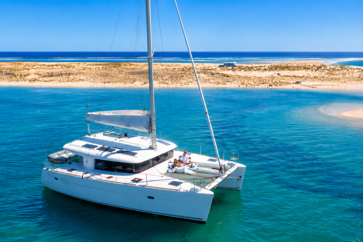 White catamaran sailing near a sandy island in clear blue waters.