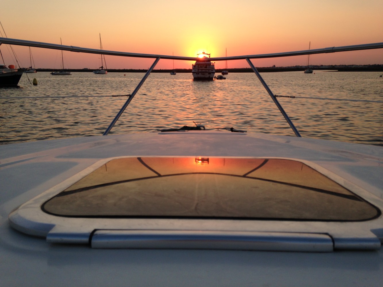 Boat deck view with sunset over calm water and anchored boats.