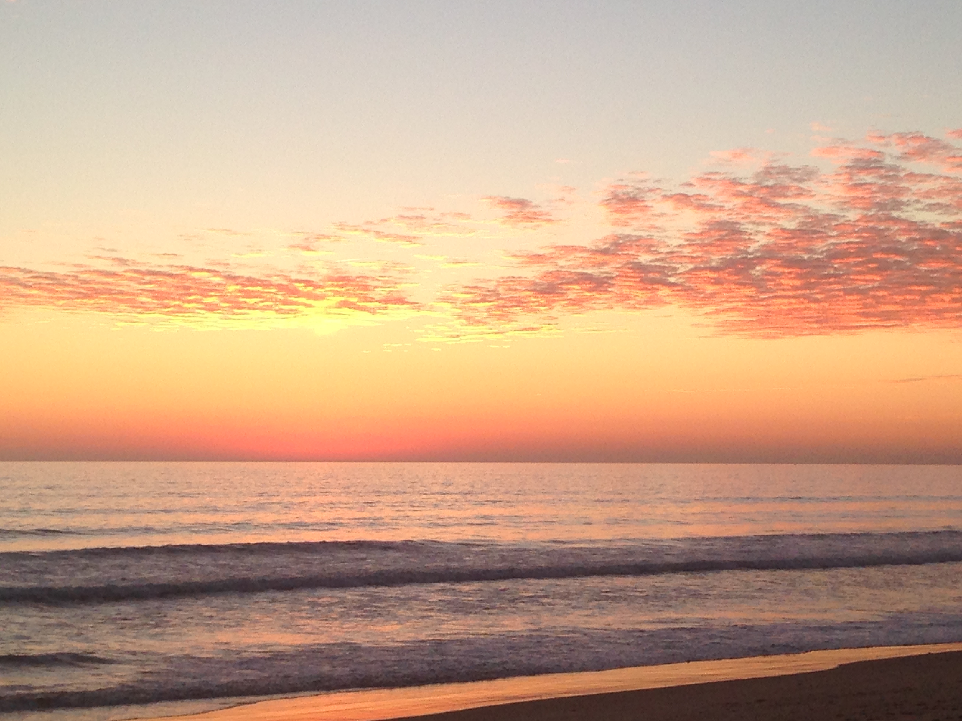 Sunset over ocean with pink and orange clouds, waves gently lapping at shore.