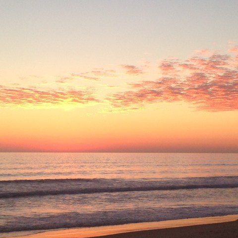Sunset over ocean with pink and orange clouds, waves gently lapping at shore.