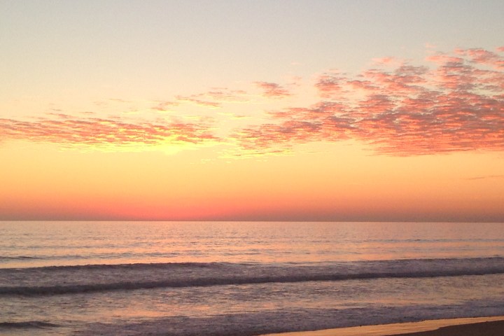 Sunset over ocean with pink and orange clouds, waves gently lapping at shore.