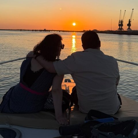 Couple on boat watching sunset over calm water with cranes in distance.