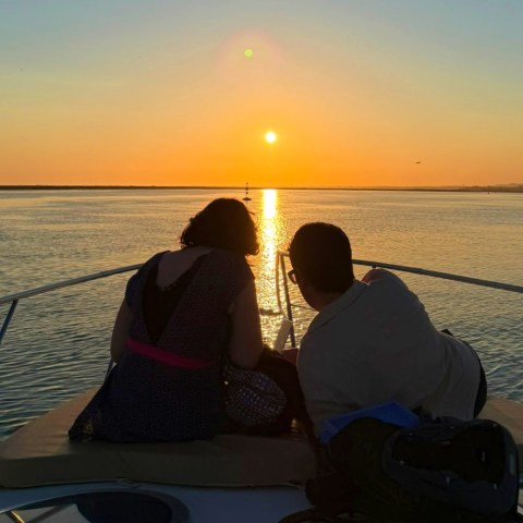 Two people sit on a boat watching sunset over water.