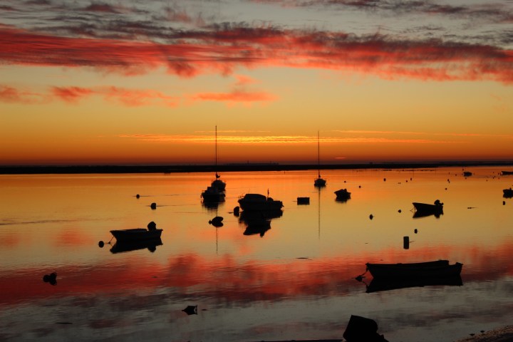 Boats silhouetted on a reflective lake at sunset with red clouds in the sky.